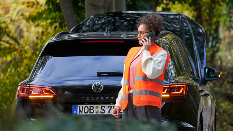 A lady in a high-visibility vest using her phone and standing next to a Volkswagen Touareg pulled over at the side of a road with the bonnet open.
