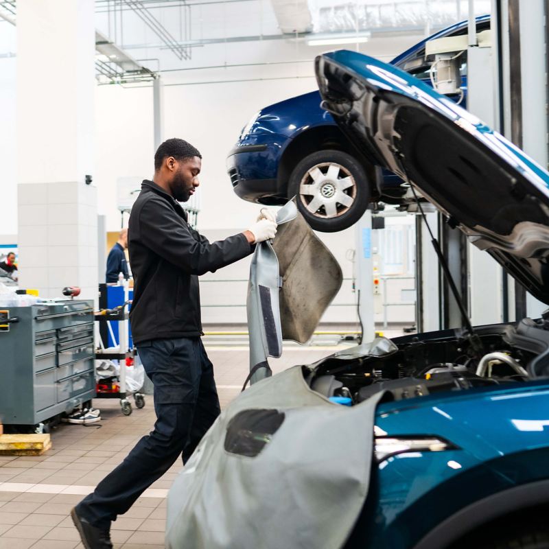 A technician standing over an open bonnet of a VW holding a car cover