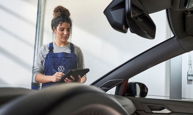 a woman wearing VW branded overalls standing next to a car, holding an ipad