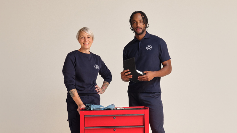 Two Volkswagen service employees standing behind a red tool cart on a plain background