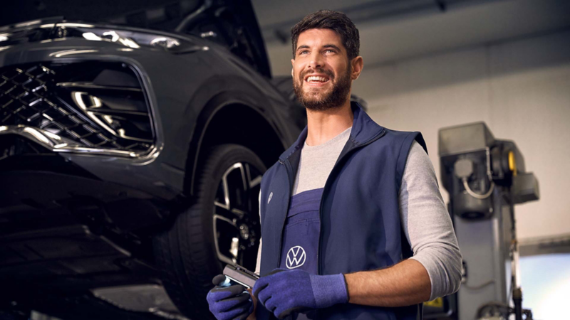 Técnico de Volkswagen sonríe junto a auto negro durante servicio de mantenimiento con herramienta en mano.