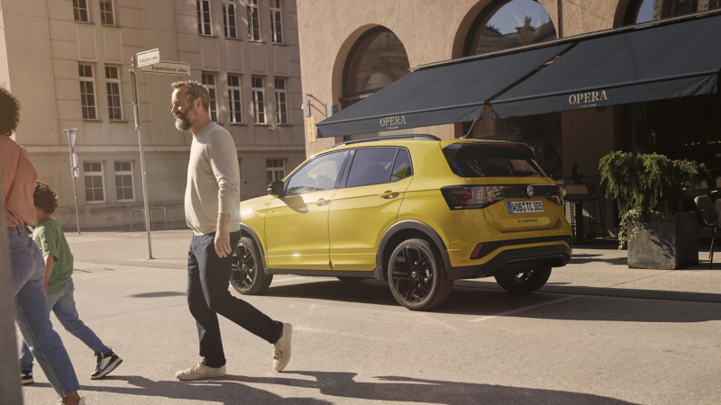 Un Volkswagen de color amarillo parado frente a una cafetería con una familia pasando delante