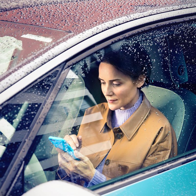 Näkymä VW ID.3:n sivuikkunasta kohti naista, joka käyttää älypuhelinta