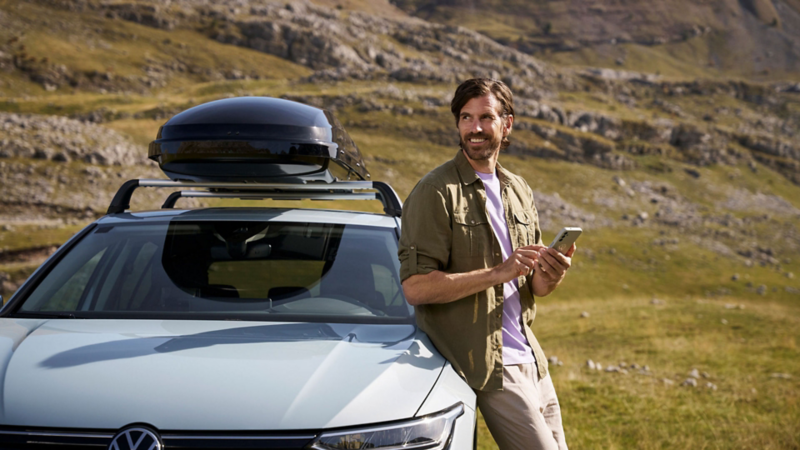 A man using a smartphone while leaning on a white Volkswagen car with a roof box in the mountains.