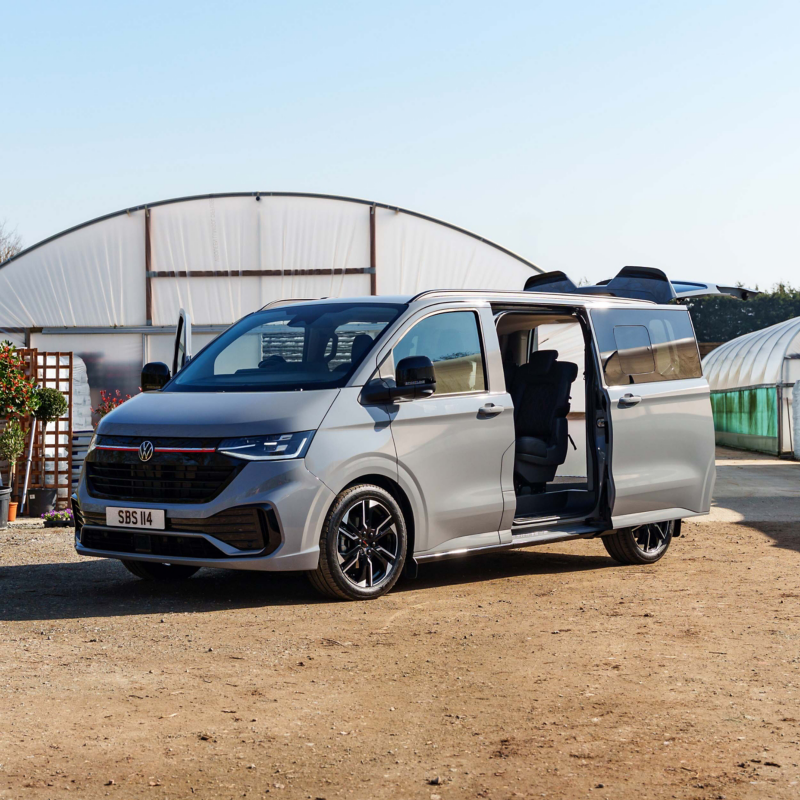 A sleek silver van with open sliding doors is parked on a sunlit dirt path. It has dark tinted windows and modern black alloy wheels, positioned near greenhouses.