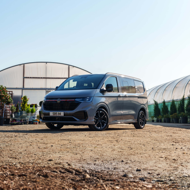 Silver van parked in front of greenhouses on a sunny day. Surrounding the van are various plants, creating a serene and relaxed atmosphere.