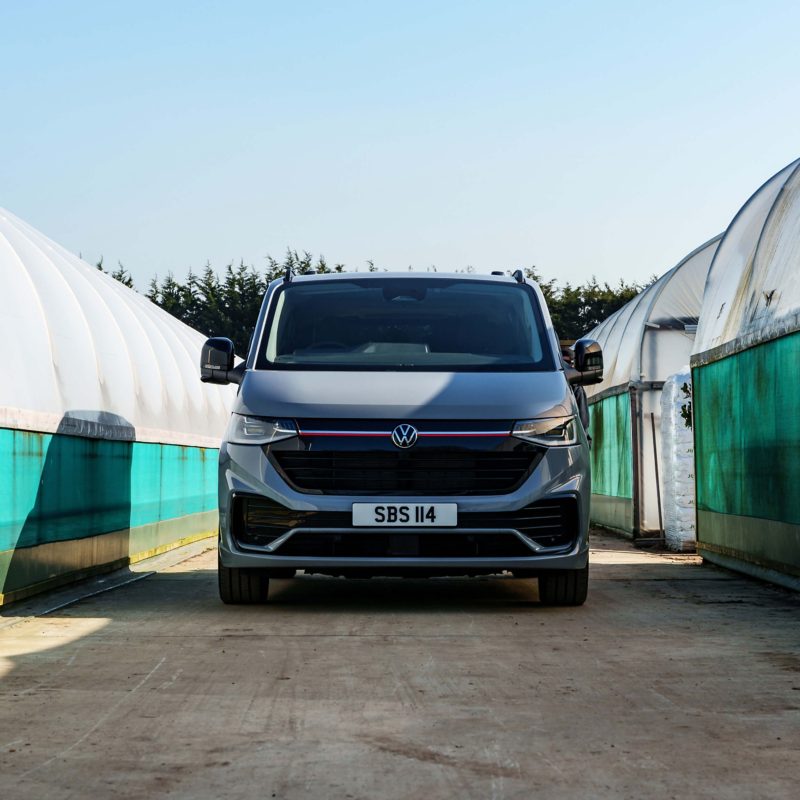 A gray Volkswagen van is parked on a dirt path between two large, translucent greenhouses under a clear blue sky, conveying calm and modernity.