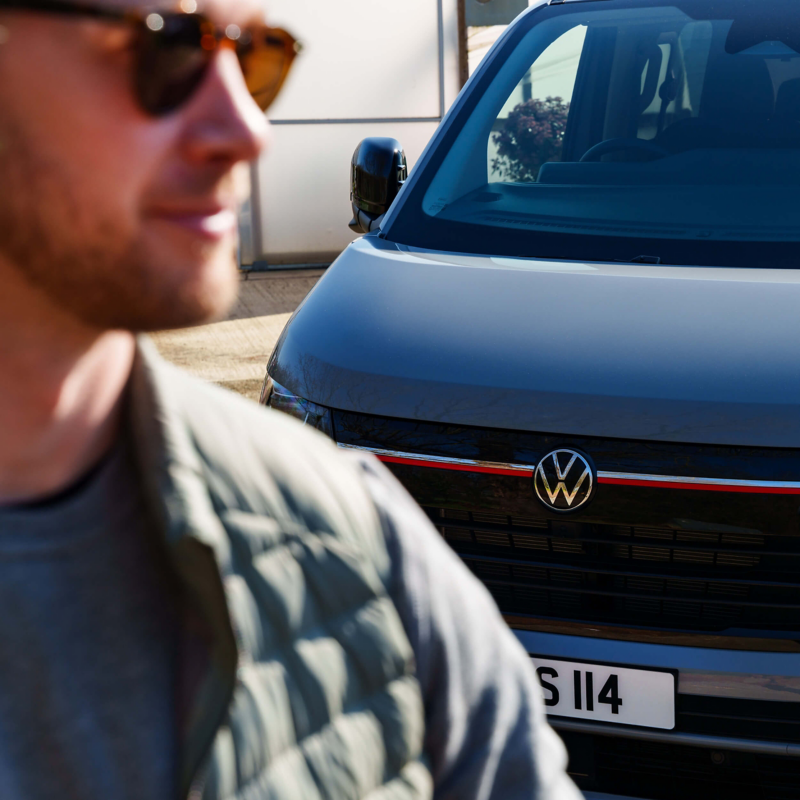 Blurred man in sunglasses and jacket in the foreground with a focused gray Volkswagen vehicle in the background, conveying a casual, sunny day.