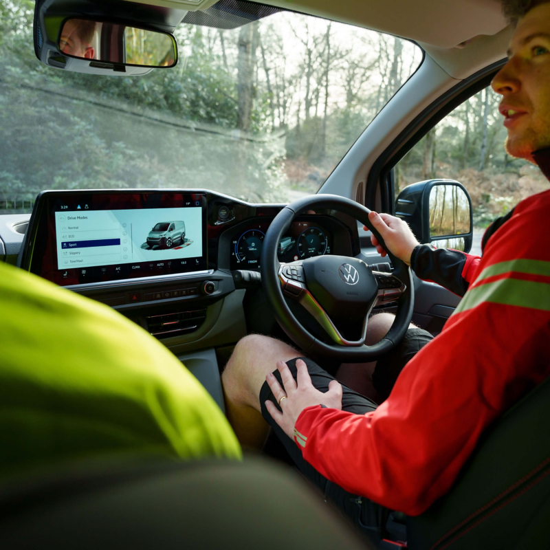 A man in a red jacket is driving a Volkswagen with a focus on the digital dashboard displaying driving modes. The mood is relaxed and modern.