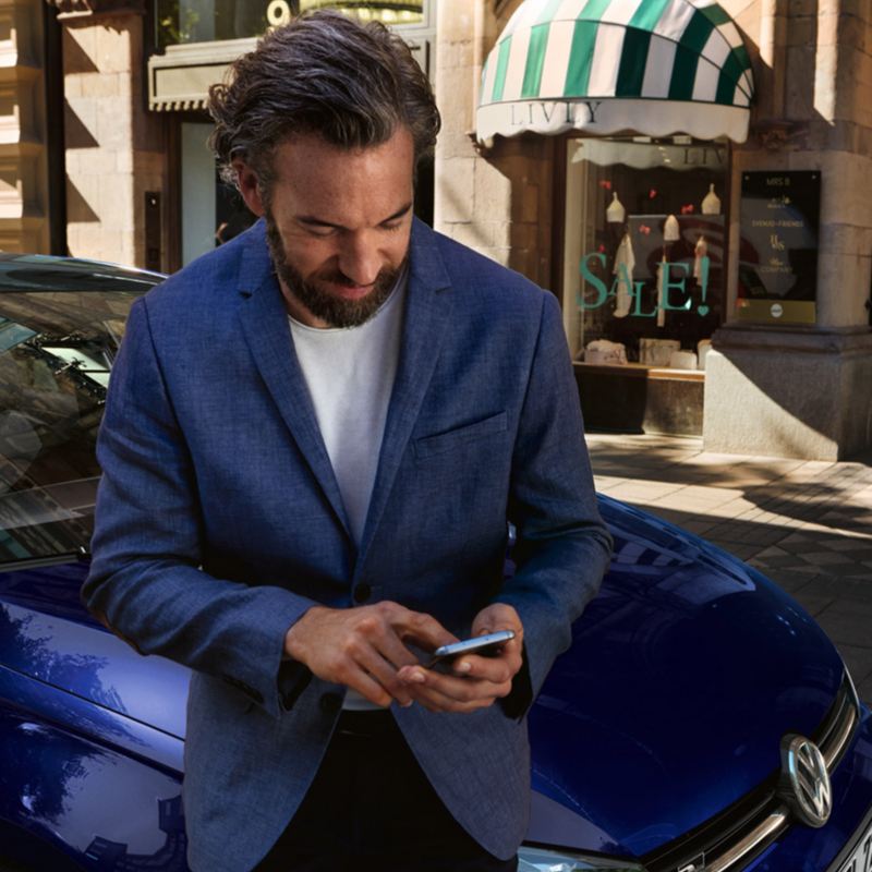 Un uomo consulta il suo smartphone appoggiato alla fiancata di un'auto Volkswagen, vista 3/4 posteriormente.