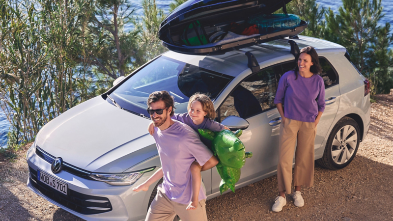  Une famille pose à côté d'une Volkswagen Golf blanche, équipée d'un coffre de toit. Le père porte son fils sur son dos, tenant un crocodile gonflable. La mère se tient à leurs côtés, et ils sourient tous les trois.