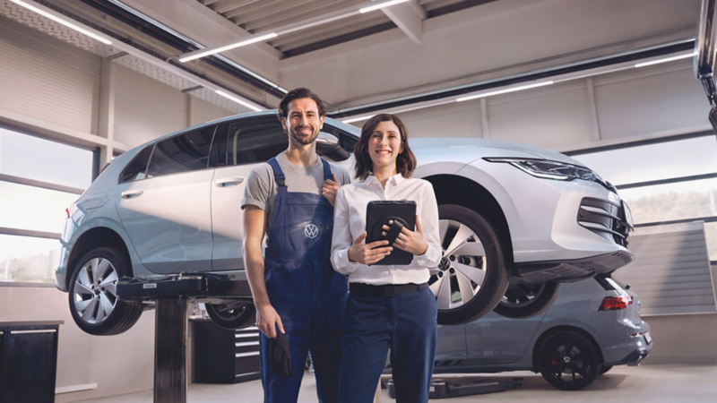 Zwei Personen in einer Werkstatt vor einem Volkswagen Golf auf einer Hebebühne, eine hält ein Tablet.