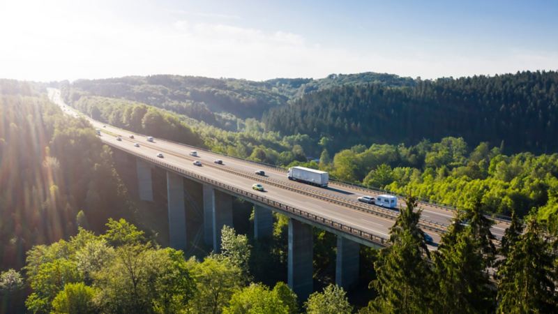 Cars and trucks drive over a bridge surrounded by mountains and forest 