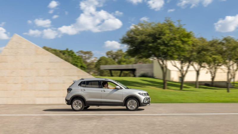 Volkswagen Taigun. Vista lateral de SUV de color plata en movimiento frente a edificio moderno.