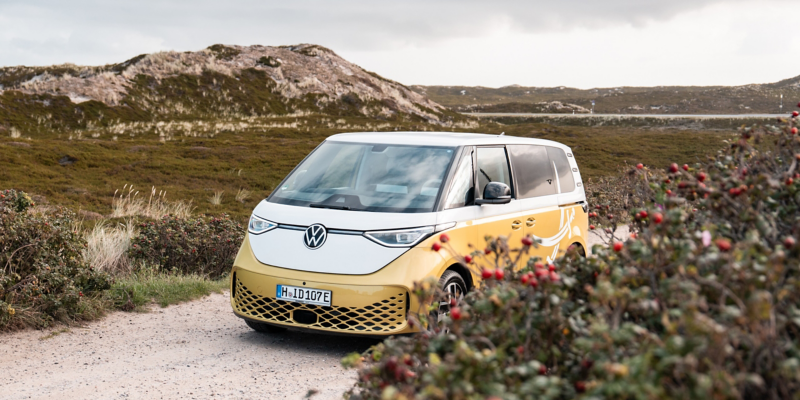 Ansicht von schräg vorn: Ein ID. Buzz Shuttle in Zweifarblackierung mit dem Logo der Sansibar auf der Fahrerseite steht auf einem Weg zwischen den Dünen auf Sylt.