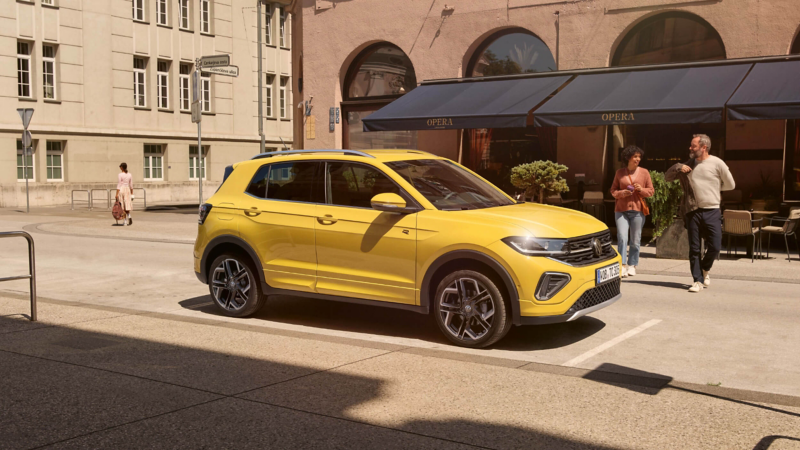 A yellow T-Cross parked in a street with two people walking towards it