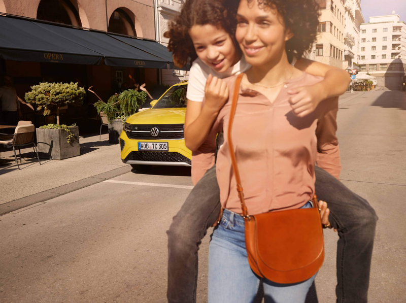 Smiling woman with a child on her back walks in sunny city street. Yellow Volkswagen T-Cross parked nearby. Warm, joyful atmosphere with urban backdrop.
