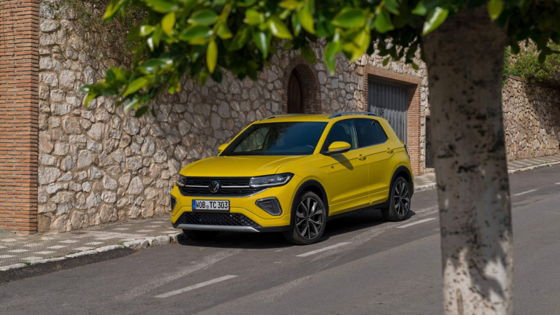 A bright yellow Volkswagen T-Cross SUV is parked next to a stone wall on a quiet street. A leafy tree partially frames the right side, conveying a relaxed, sunny setting.
