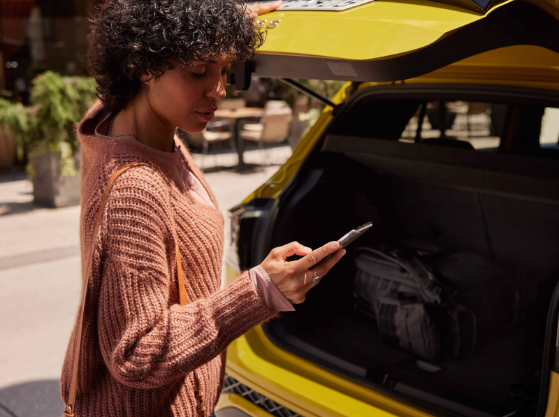 A woman with curly hair, wearing a pink sweater, stands by an open yellow boot of a T-Cross, looking at her phone. The scene is outdoors and sunny.