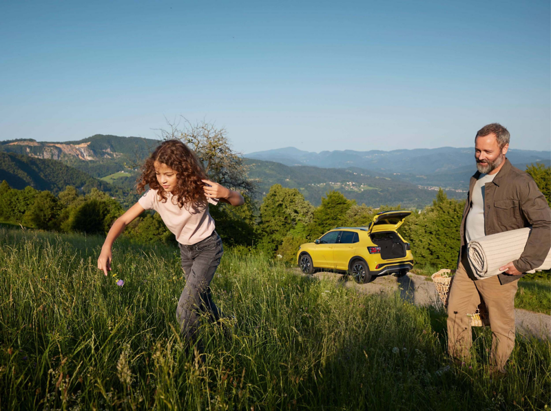 A man and girl walking up a grassy hill with a yellow T-Cross in the background