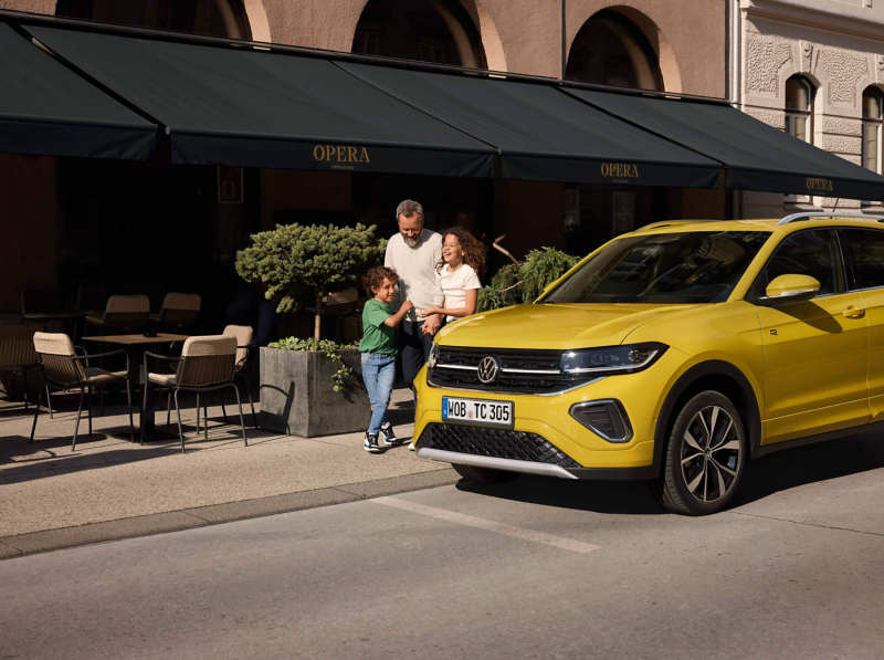A man and two children standing in the street outside of a restaurant, with a yellow VW T-Cross next to them
