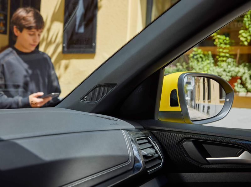 View from inside a T-Cross shows the dashboard and side mirror with a reflection of a yellow vehicle. A person with a phone stands outside, blurred in focus.