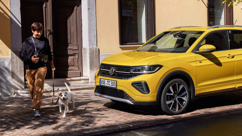 A person walks a happy dog on a cobblestone street beside a bright yellow Volkswagen Volkswagen T-Cross SUV. The scene is sunny, conveying a cheerful, relaxed mood.