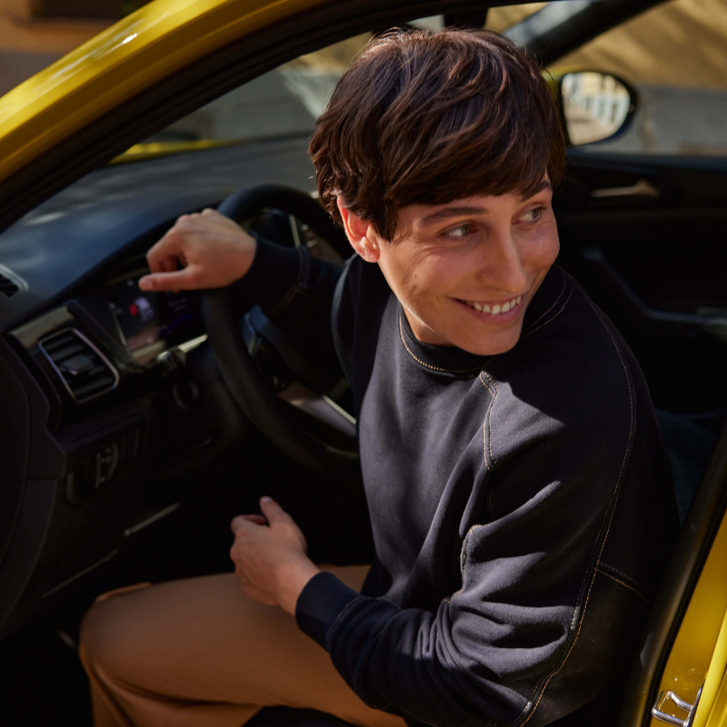 Smiling person sits in a yellow T-Cross with the door open, wearing a black sweater and tan trousers. Sunlight filters in, creating a warm, cheerful atmosphere.