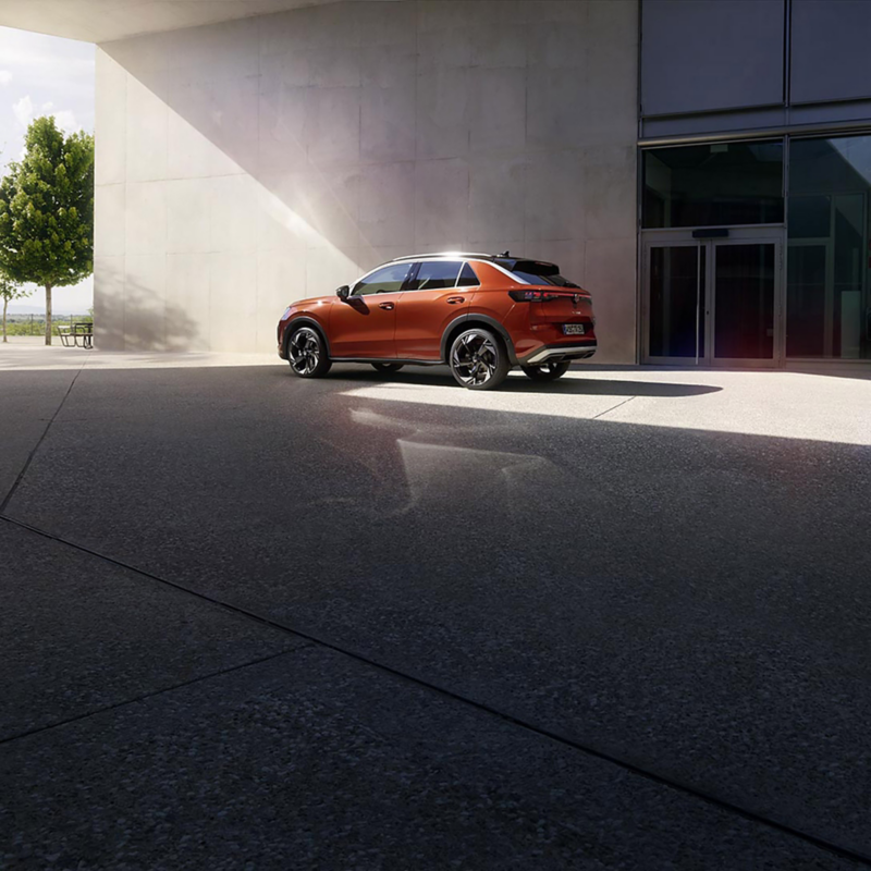 A sleek orange Volkswagen T-Roc SUV is parked outside a modern building, bathed in sunlight. A leafy tree casts a shadow on the pavement, conveying a serene urban scene.