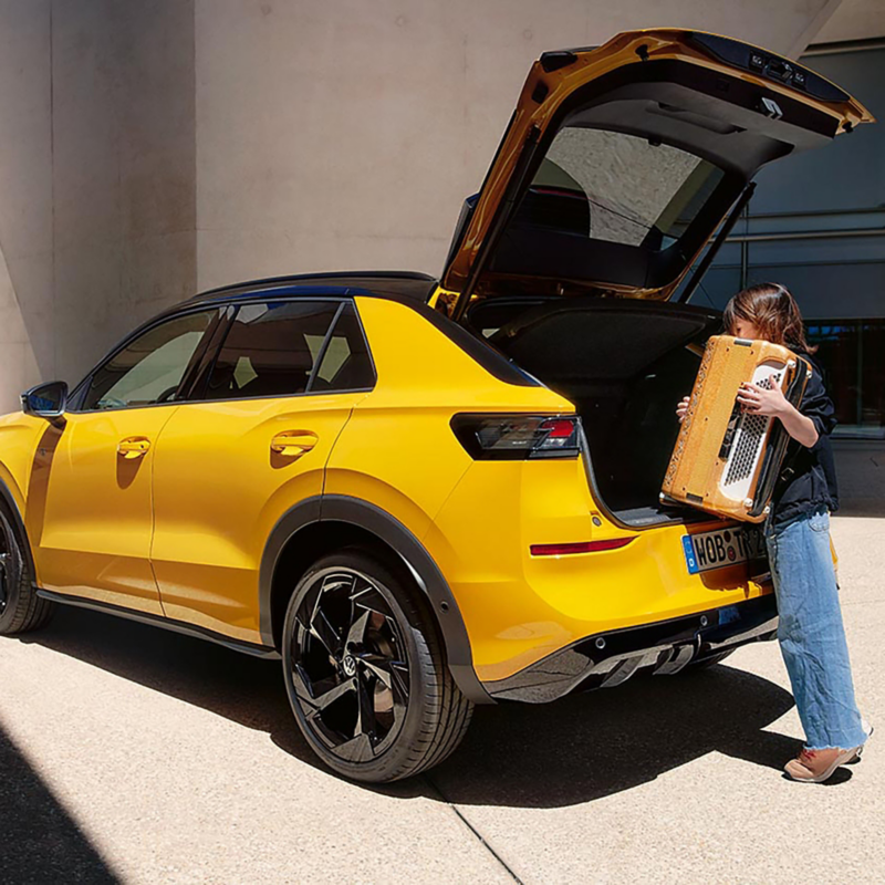 Yellow T-Roc SUV with the boot open on a sunny day. A person stands behind, lifting an accordion from the vehicle, conveying a casual and lively atmosphere.