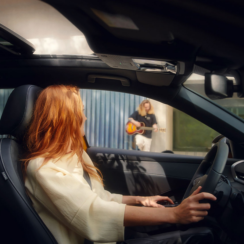 A woman with red hair sits in a T-Roc, smiling as she looks out the window at a person playing a guitar. The scene conveys a warm, relaxed atmosphere.