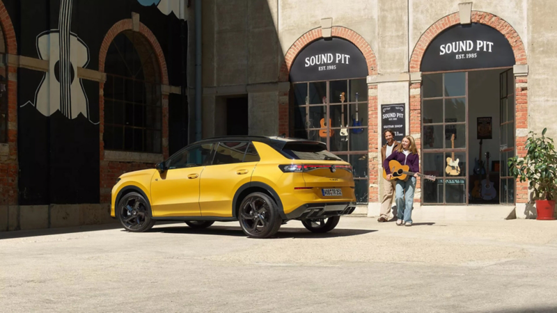A vibrant yellow Volkswagen T-Roc SUV is parked in a sunlit courtyard outside a shop called "Sound Pit." Two people with a guitar stand nearby, exuding a cheerful vibe.
