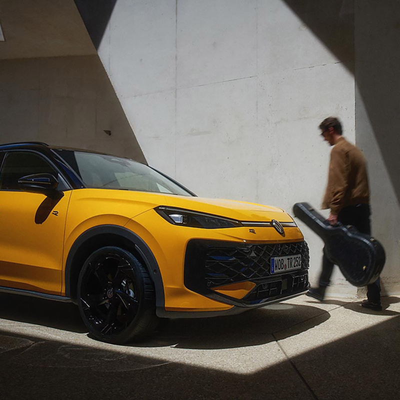 A sleek yellow Volkswagen T-Roc is parked in a sunlit area against a concrete wall. A person walks by holding a guitar case, creating a modern, dynamic scene.