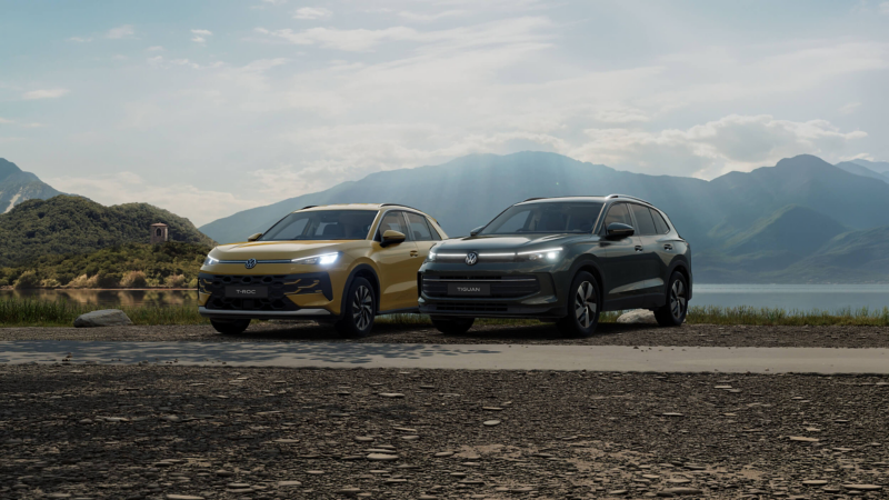 Two Volkswagen SUVs, a T-Roc and a Tiguan, are parked on a rocky lakeside road beneath misty mountains and a cloudy sky, conveying a calm, adventurous mood.