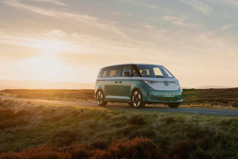 Front three-quarter view of a two-tone green and white Volkswagen ID. Buzz electric van parked on a rural road at sunset.