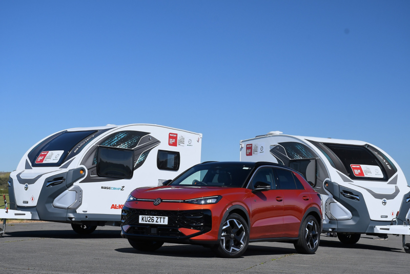 Front three-quarter view of an orange Volkswagen SUV parked between two modern white Swift Basecamp caravans under a clear blue sky.