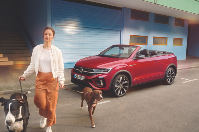 Frau führt zwei Hunde an der Leine vor einem roten VW T-Roc Cabriolet mit geöffnetem Verdeck