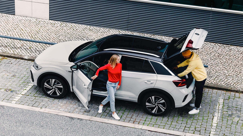 An aerial view of a woman getting out of a white VW T-Roc, and a man accessing the boot