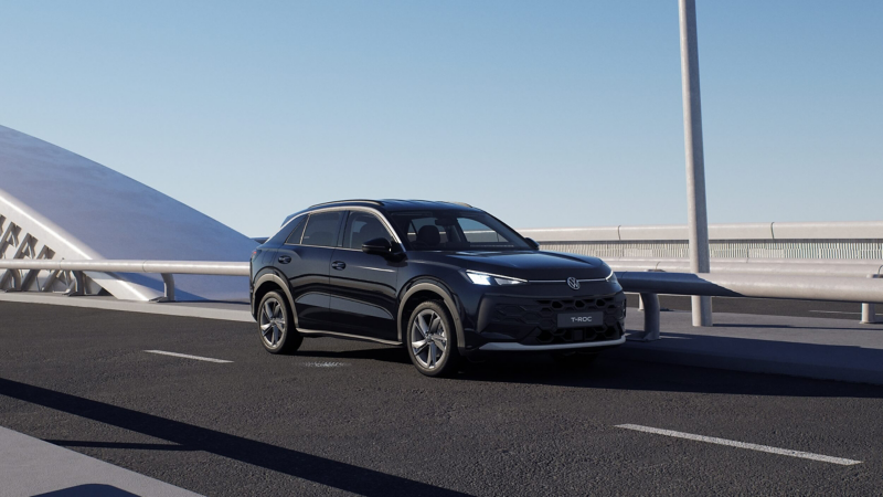 A dark VW T-Roc Match is driving across a bridge with a sunny blue sky as the background.