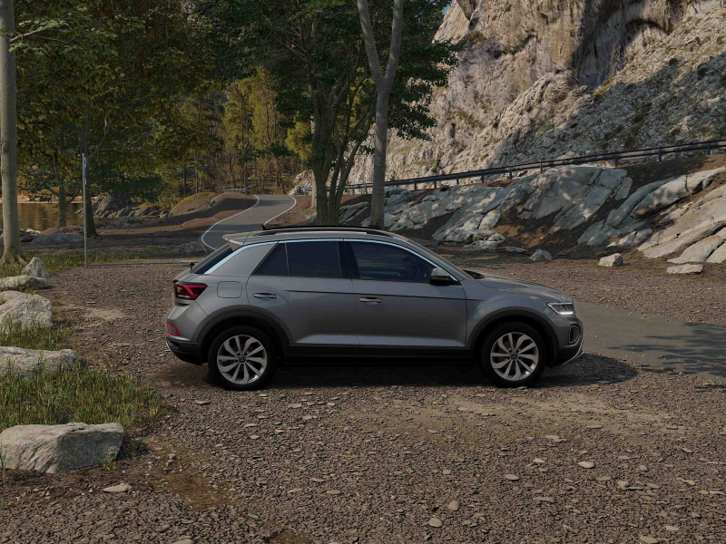 A silver Volkswagen T-Roc SUV is parked on a gravel lot surrounded by rocky hills and trees, under a clear sky, conveying a sense of adventure and tranquility.