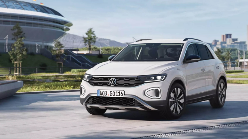 A white Volkswagen T-Roc SUV parked on a paved plaza with a futuristic stadium building in the background.