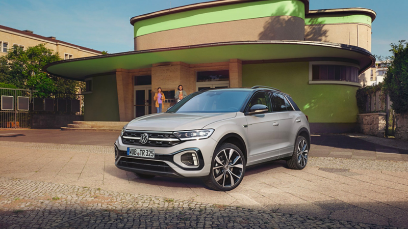 VW T-Roc gris garé devant une maison avec des personnes qui s'approchent à l'arrière