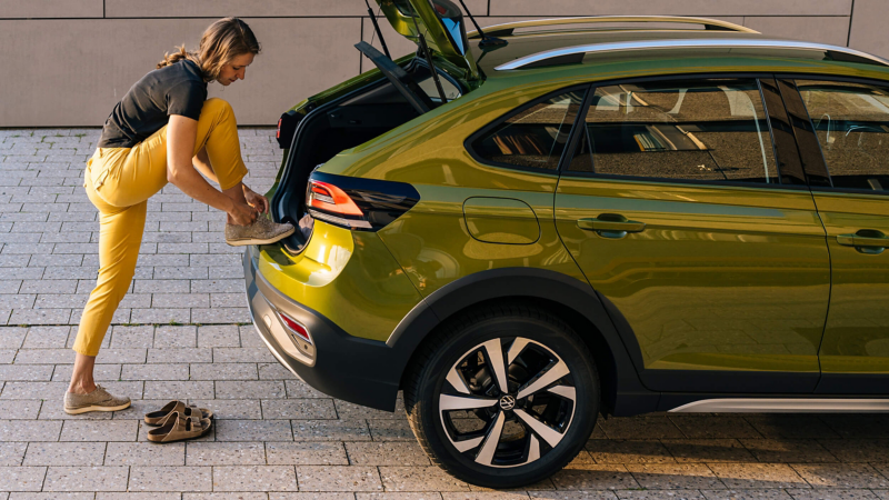 A woman tying her shoelace, while leaning on the rear section of a VW Taigo. 