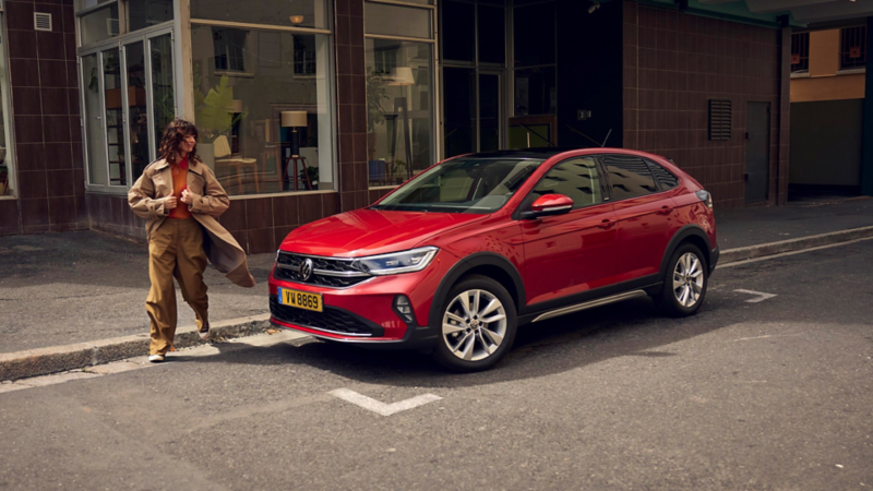 Une VW Taigo rouge est garée devant un magasin, tandis qu'une femme passe à côté.