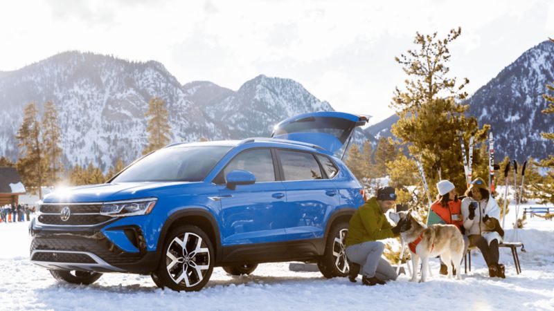 Deux femmes discutent et un homme est avec un chien près d’un VWUS Taos 2022 de Volkswagen couleur bleuet dans une station de ski.