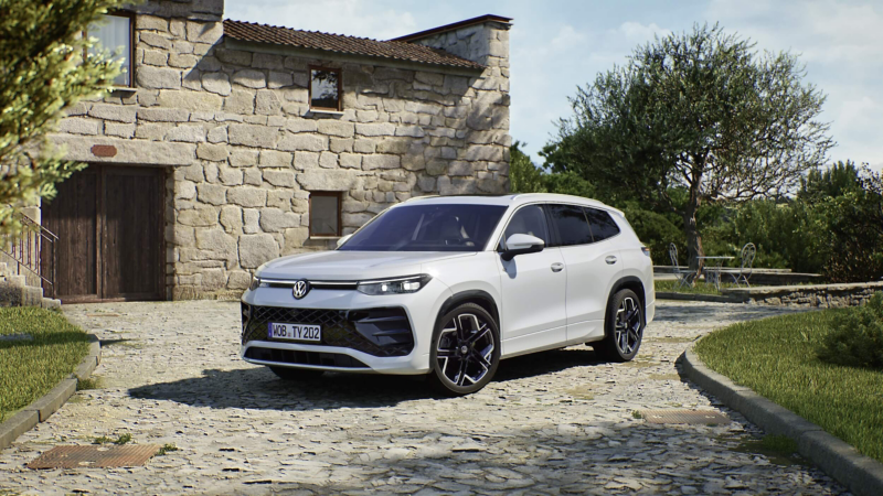 A white Volkswagen Tayron SUV with its headlights on is parked on a cobblestone street, surrounded by lush green trees and a modern building, creating a serene and modern ambiance.