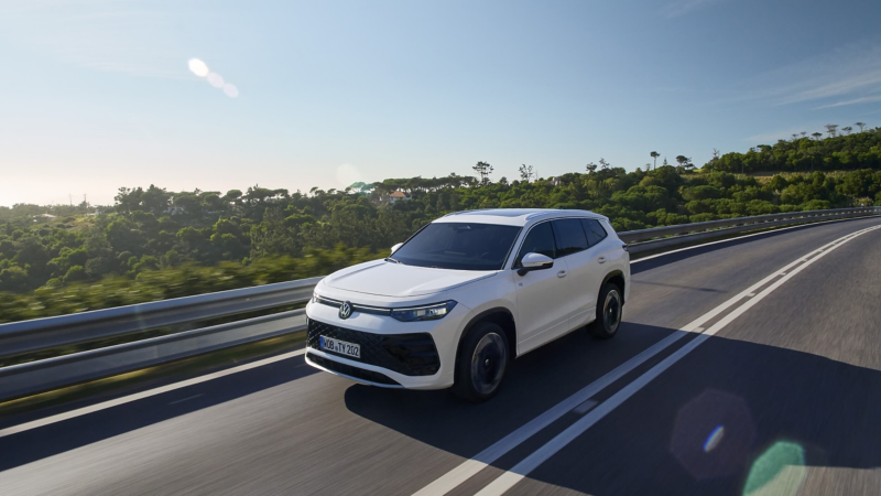 A white Volkswagen Tayron SUV travels on a sunny, open road bordered by lush greenery. The scene conveys a sense of freedom and adventure, with a clear blue sky above.