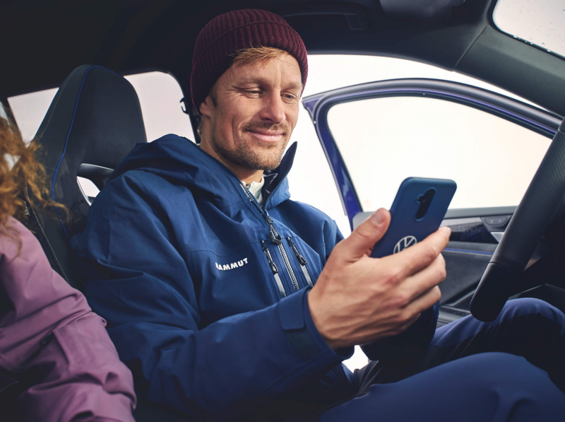 A man in a blue jacket and red beanie smiles while looking at his phone inside his Volkswagen Tayron. The car door is open, and the atmosphere is relaxed.