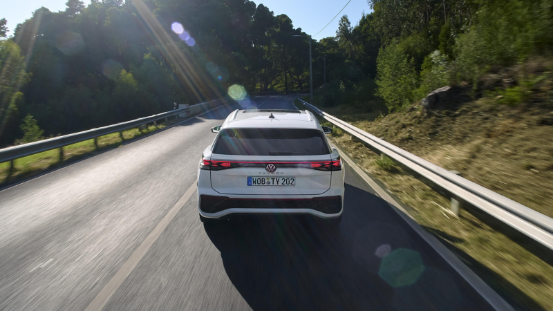 A white Volkswagen Tayron car drives down a sunlit road, lined with metal guardrails and green trees, exuding a sense of speed and freedom on a clear day.