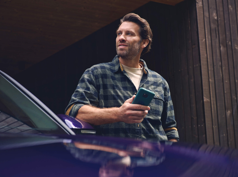 A man in a checkered shirt holds a smartphone, leaning on a Tayron with a contemplative expression. The background is a wooden building exterior.